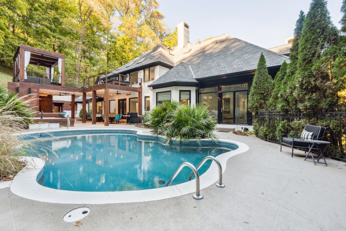 A kidney-shaped swimming pool is situated in the backyard of a large house, with a pergola and deck visible in the background, all surrounded by lush greenery.