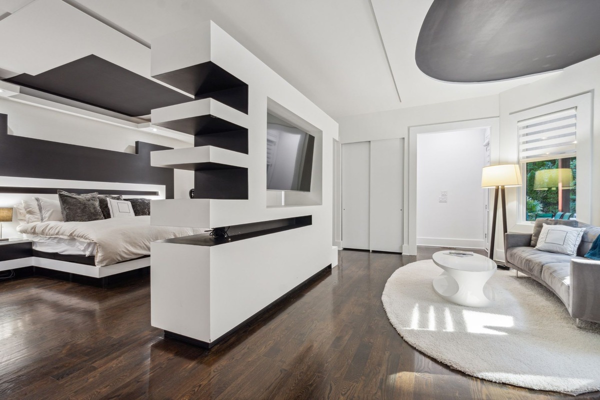 A modern bedroom features a large, built-in entertainment center separating the sleeping area from a sitting area with a curved sofa and a white coffee table on a fluffy rug.