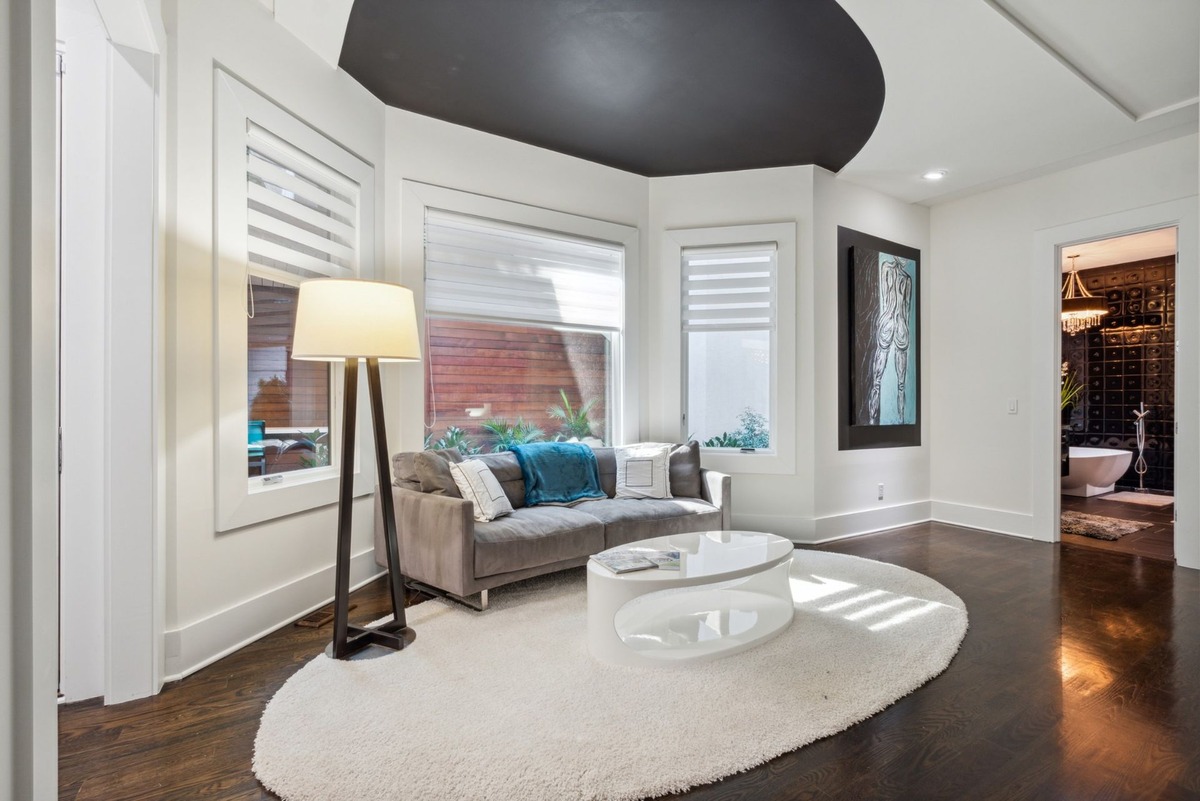 A modern living room features a gray sofa, a white coffee table on a large rug, and a floor-to-ceiling window with window shades, all set against dark hardwood floors and a partially visible bathroom doorway.