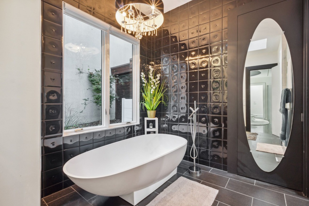 A modern bathroom features a freestanding white bathtub, dark-colored tile walls, a large oval mirror, and a chandelier above.