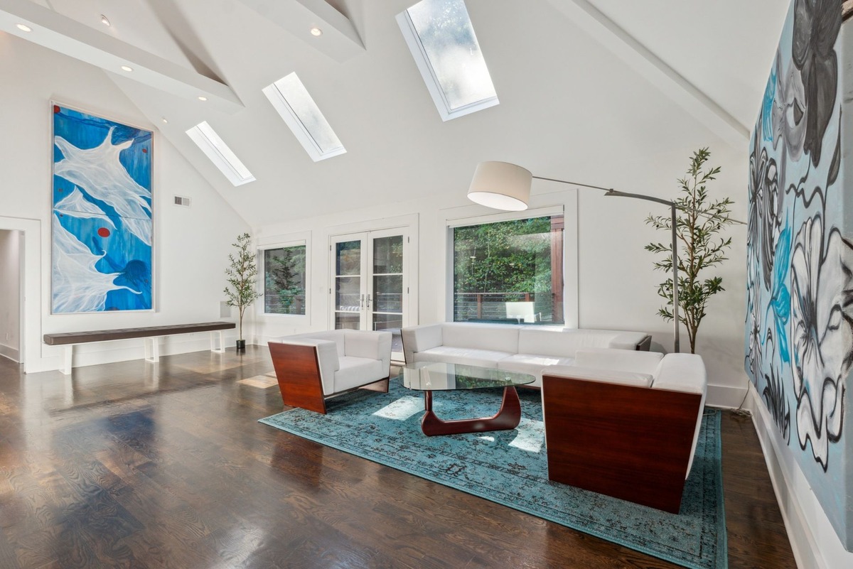 A bright, airy living room features hardwood floors, skylights, white walls, and modern white couches with wooden accents, centered around a large abstract painting.