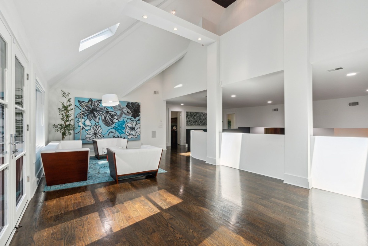 A spacious, bright living area features hardwood floors, white walls, and two white couches with wooden accents, centered around a large piece of floral artwork.