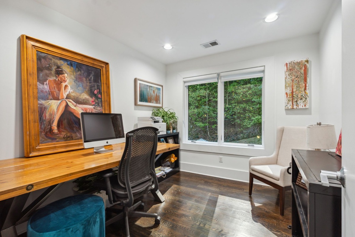 A home office features a large wooden desk with a computer, a large painting of a woman, and a comfortable off-white chair near a window overlooking trees.