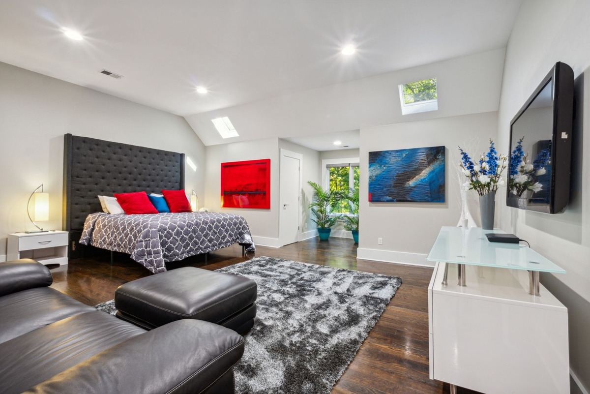 A master bedroom with a large, tufted headboard bed, dark leather ottomans, and modern artwork is shown, with a flat-screen TV and white media console visible in the foreground.