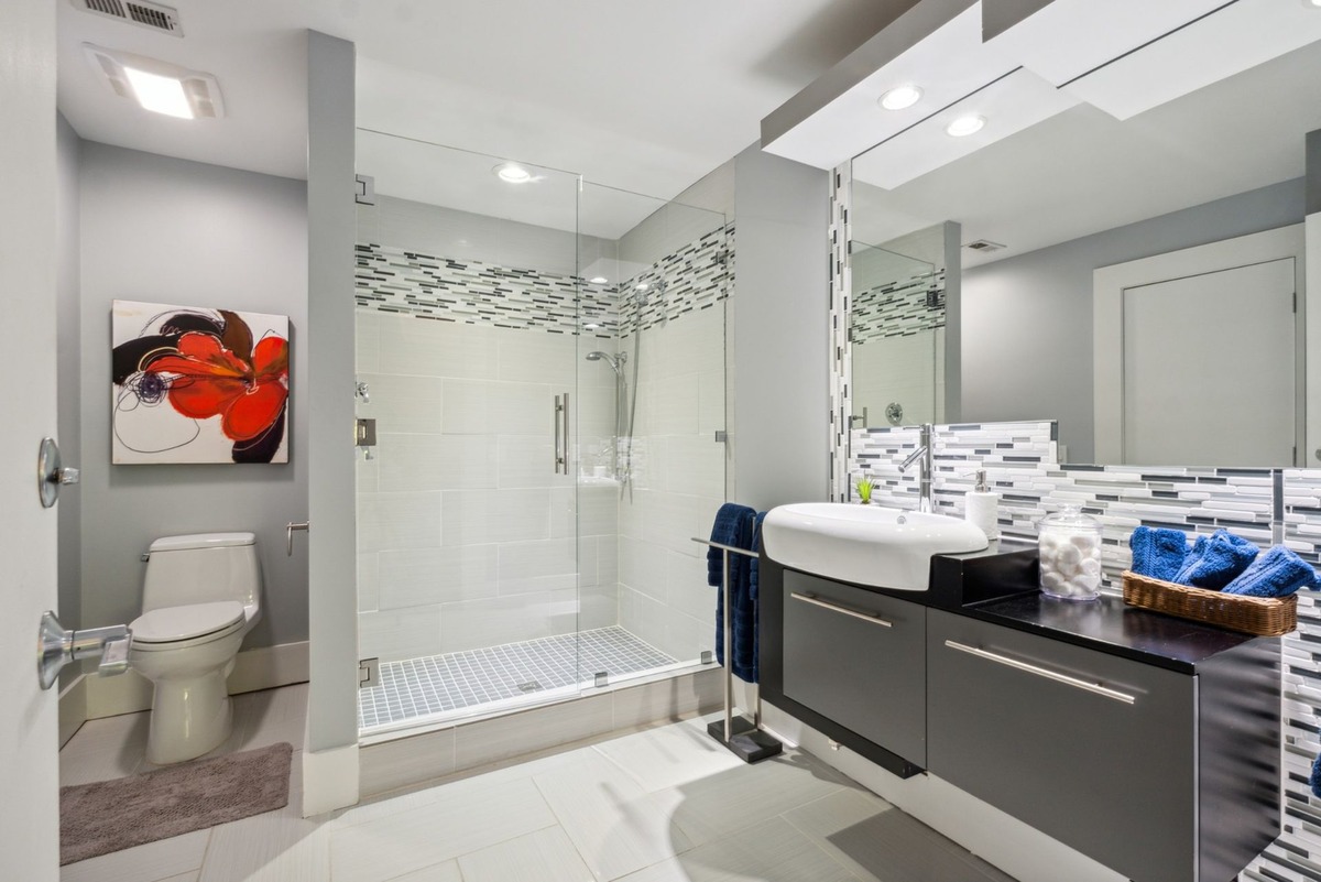 A modern bathroom features a glass-enclosed shower, a floating vanity with a vessel sink, mosaic tile accents, and a piece of abstract art.