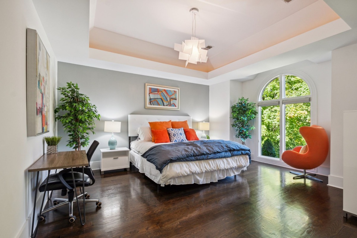 A spacious master bedroom features a king-size bed with orange and gray bedding, a dark wood floor, a large window with a view of trees, and an orange egg chair.