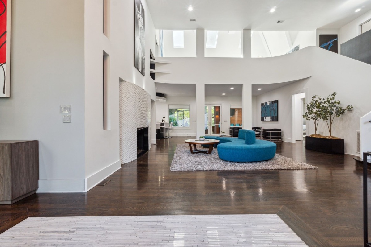 A spacious, modern living room features high ceilings, dark hardwood floors, a large blue curved sofa, and a white tiled fireplace wall.