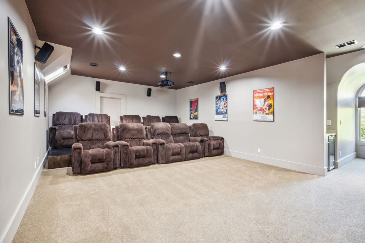 A home theater room features a row of brown reclining chairs, movie posters on the walls, and a dark brown ceiling with recessed lighting.