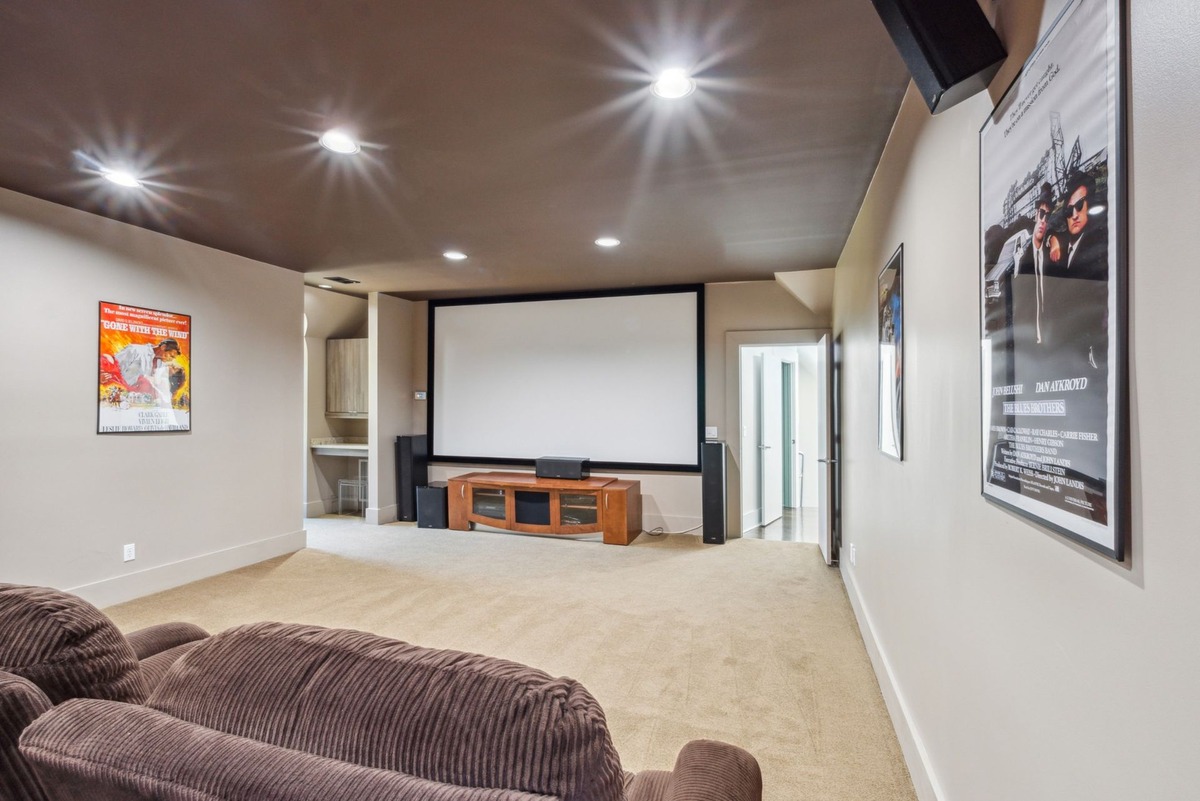 A home theater room with a large projection screen, a media console, and brown corduroy seating is shown, with movie posters adorning the walls.