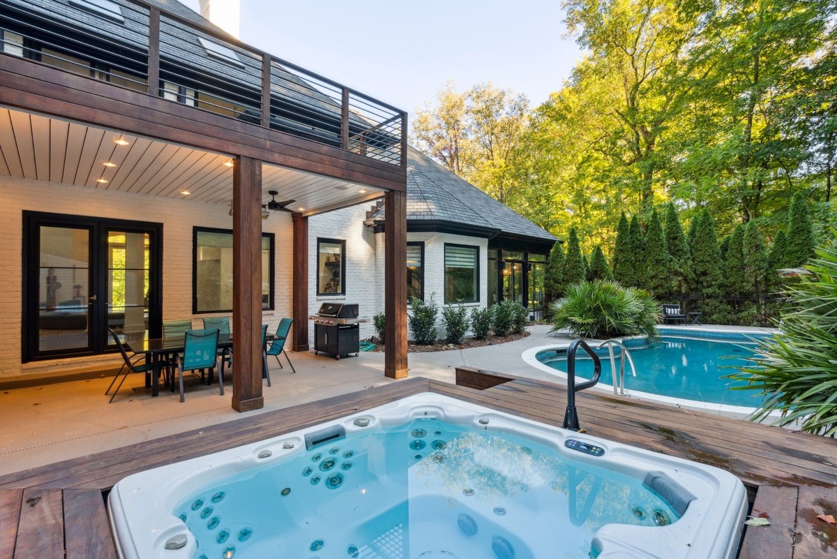 A hot tub sits on a deck overlooking a swimming pool and a modern house nestled in a wooded area.