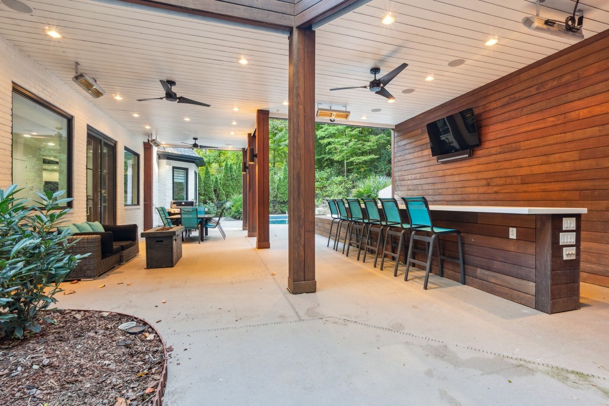 An outdoor patio area with a covered section features a long bar with teal stools, ceiling fans, and views of a pool and wooded area beyond.