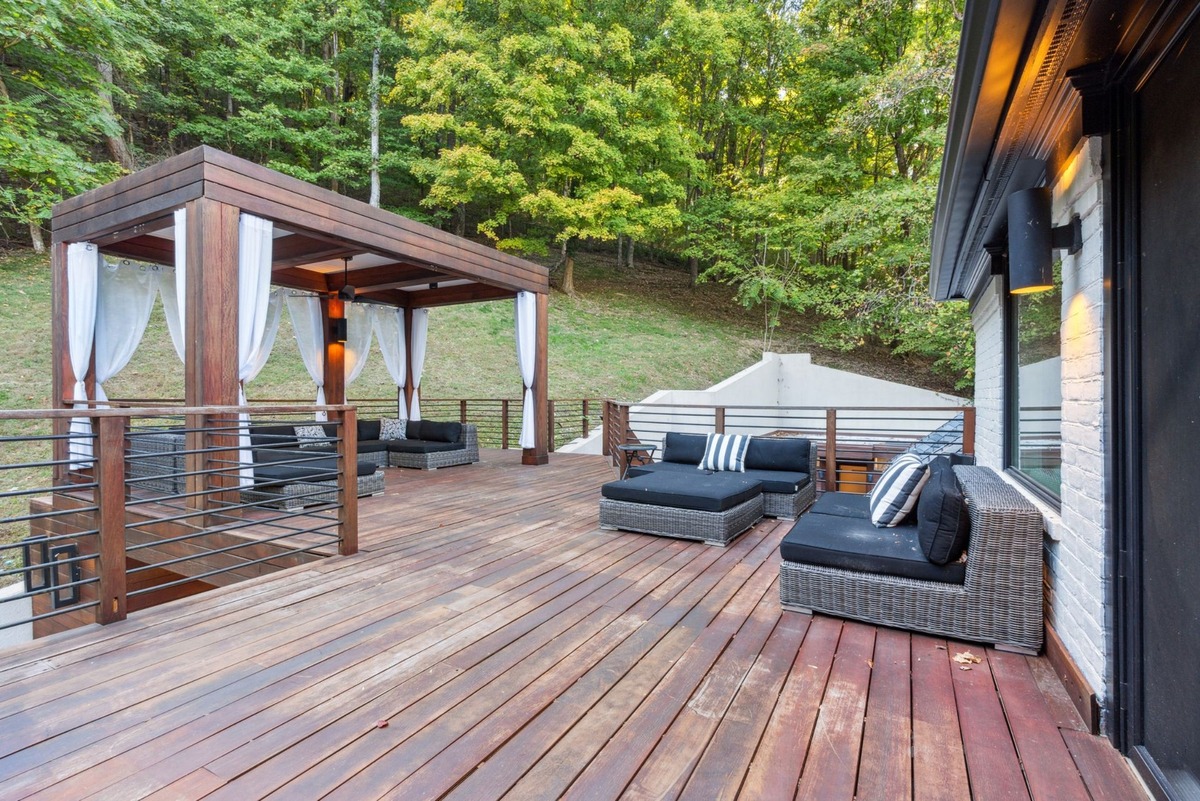 A large wooden deck overlooks a wooded hillside and features a pergola with white curtains, several wicker seating areas, and a modern home in the background.