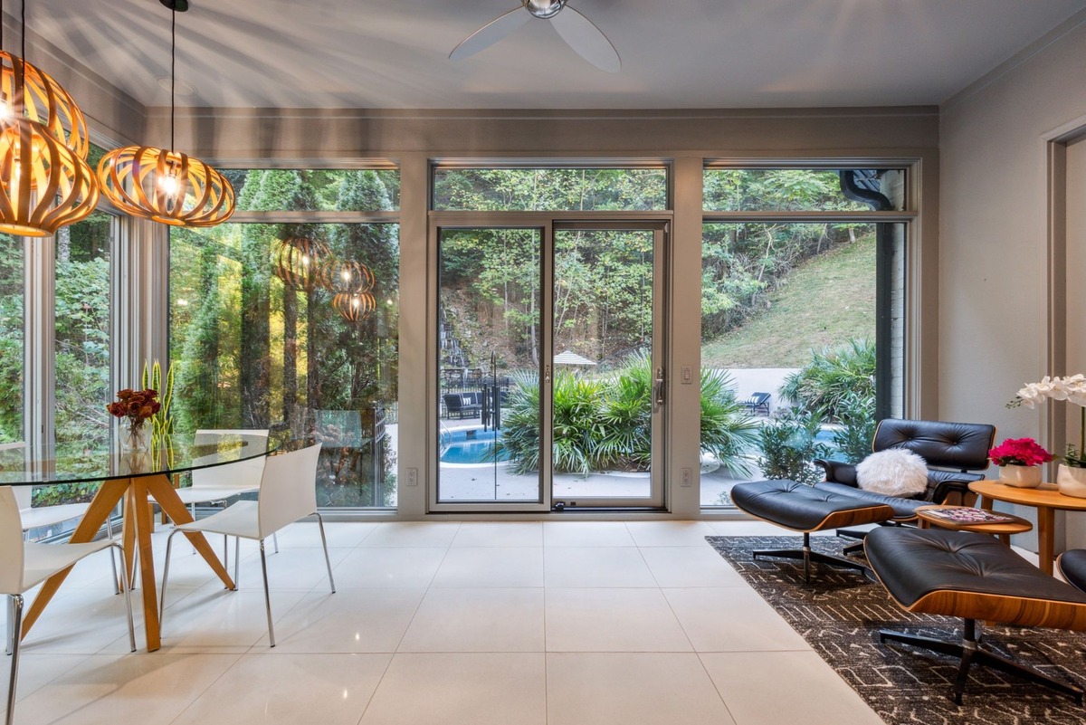 A sunroom with large windows overlooking a wooded area and a pool, features a glass dining table with white chairs, and a modern leather lounge chair and ottoman.