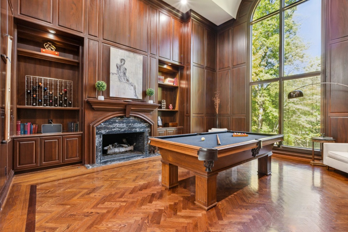 A game room with dark wood paneling, a large window, a fireplace, built-in shelving, and a pool table in the center is shown.