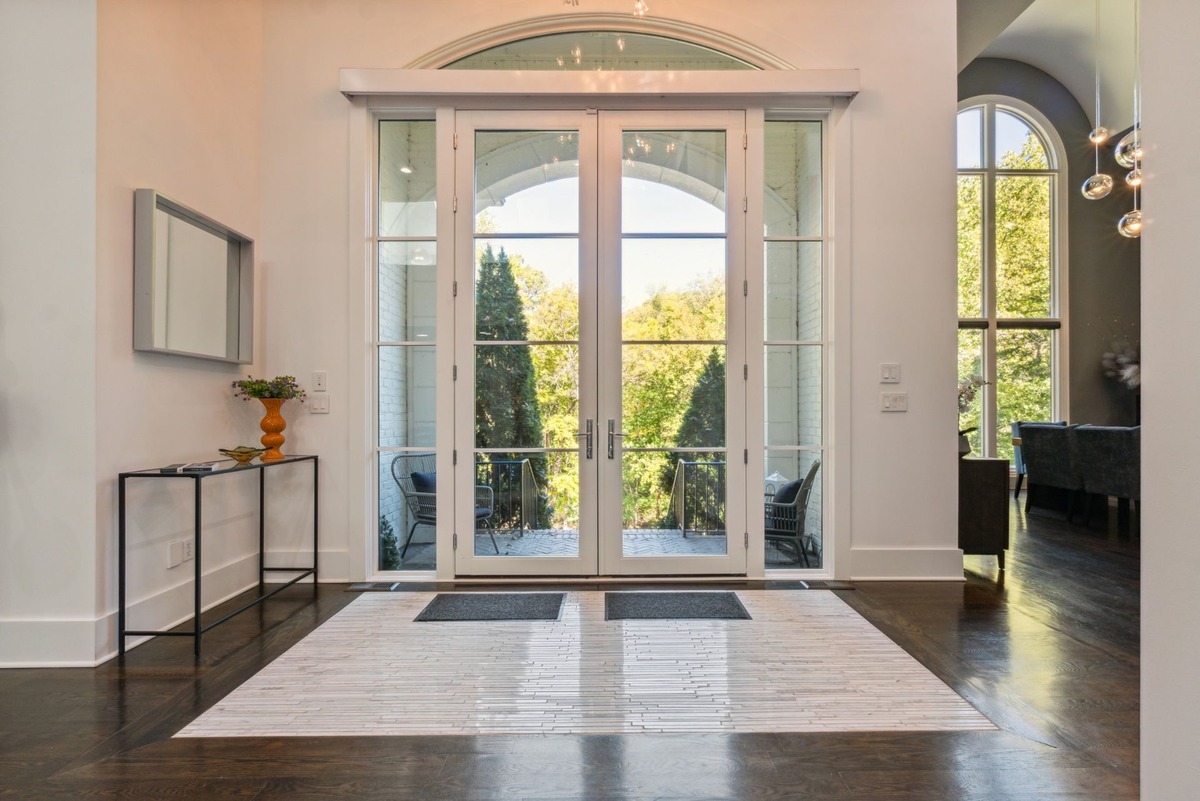 A bright entryway features a large pair of French doors opening onto a patio, a long black console table, and a light-colored rug on dark hardwood floors.