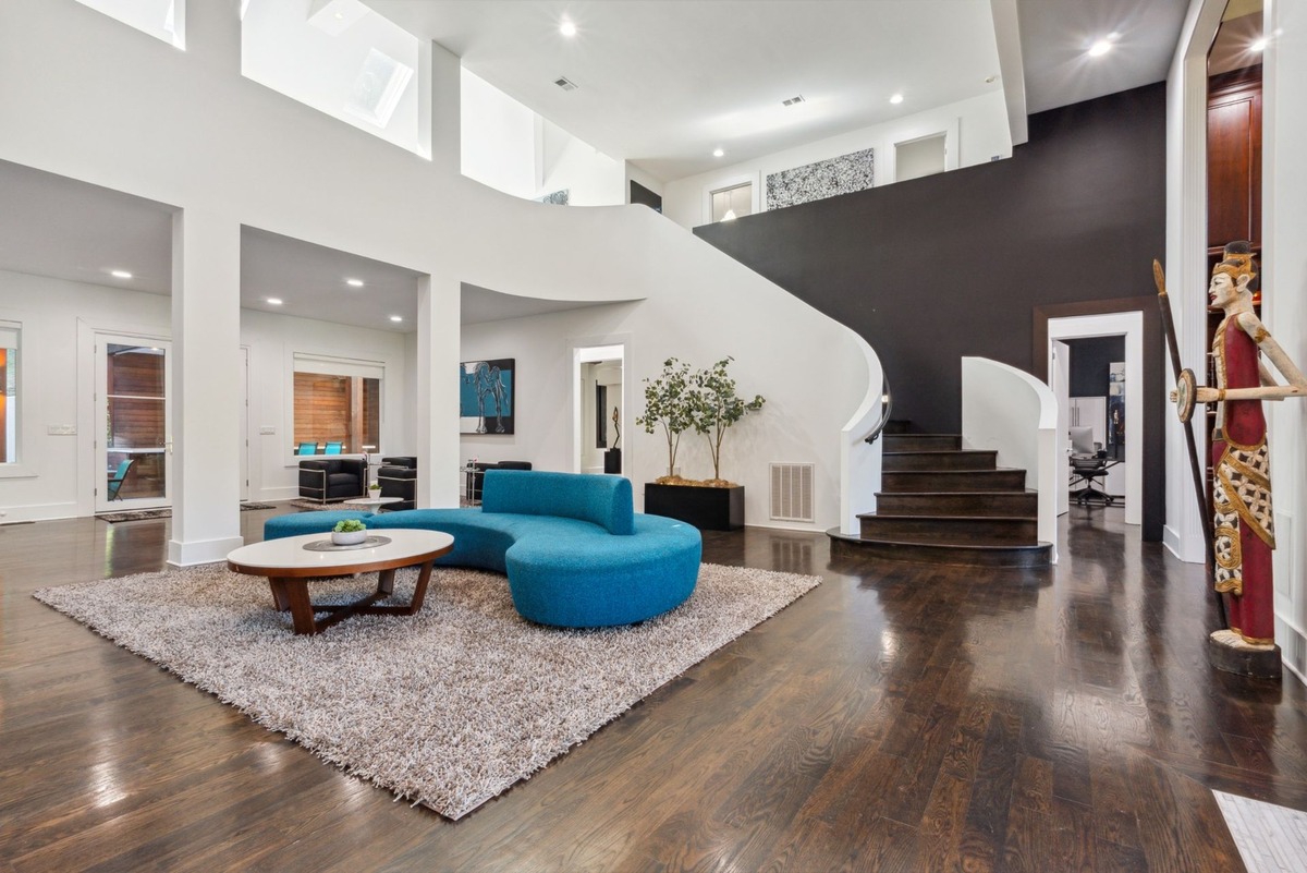 A modern, open-plan living area features a curved blue sofa, a circular coffee table, dark hardwood floors, and a curved staircase leading to an upper level.