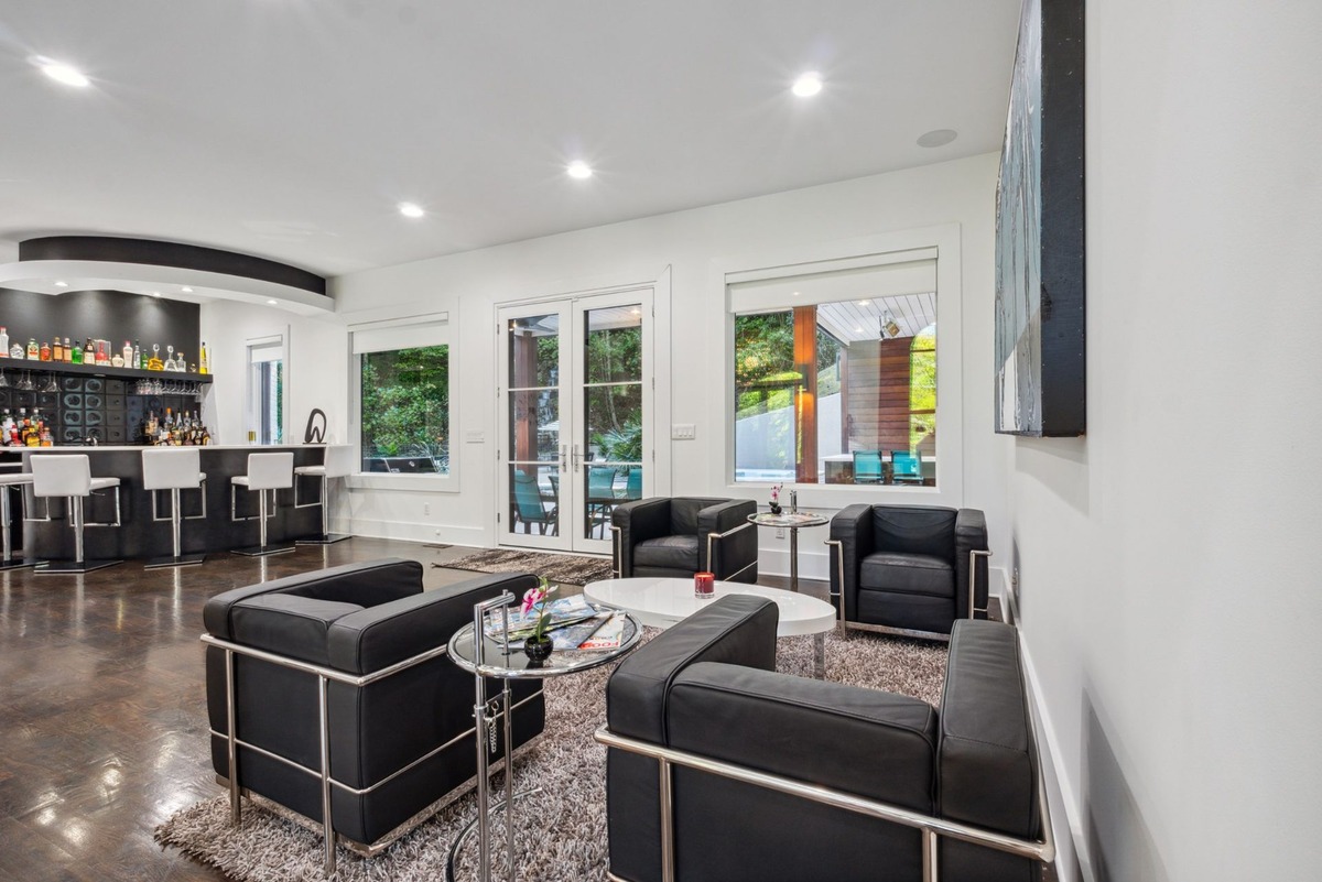 A modern living area features dark leather armchairs arranged around a low coffee table, with a home bar visible in the background and large windows offering a view of the outdoors.