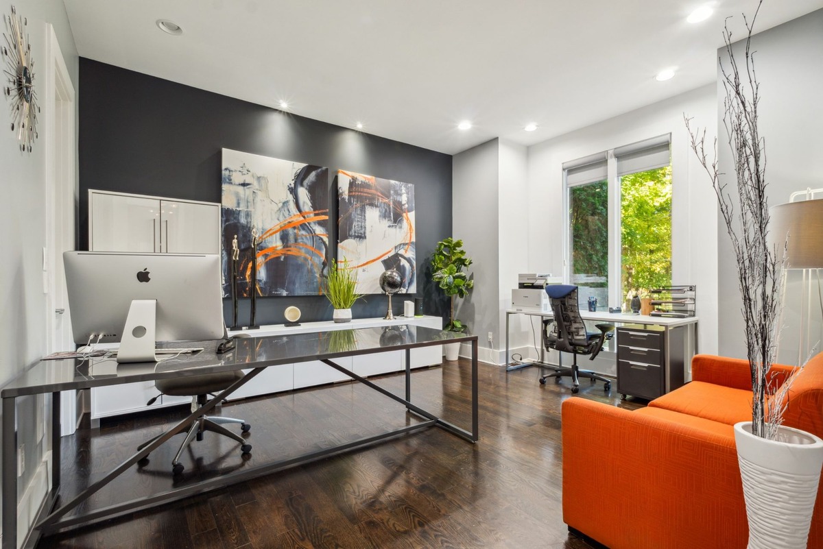 A modern home office features a large glass-topped desk with an Apple computer, an orange couch, and two large pieces of abstract art on a dark wall.