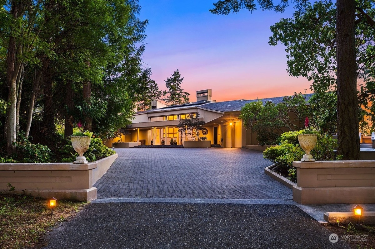 Large, luxurious home with a long driveway sits on a property surrounded by lush greenery at dusk.