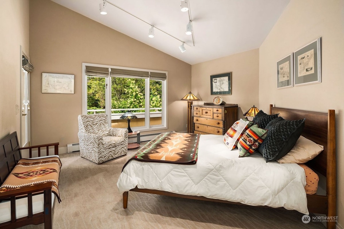 Bedroom with a king-size bed, patterned throw, and a view of trees from a large window features a dresser and a comfortable armchair.