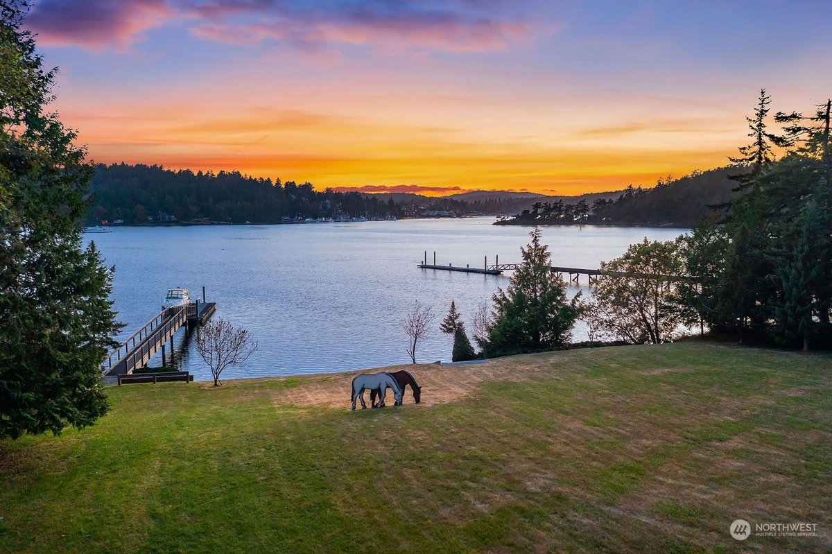 Two horses graze peacefully in a field overlooking a serene waterfront scene at sunset, with boats docked at piers and a picturesque landscape in the background.