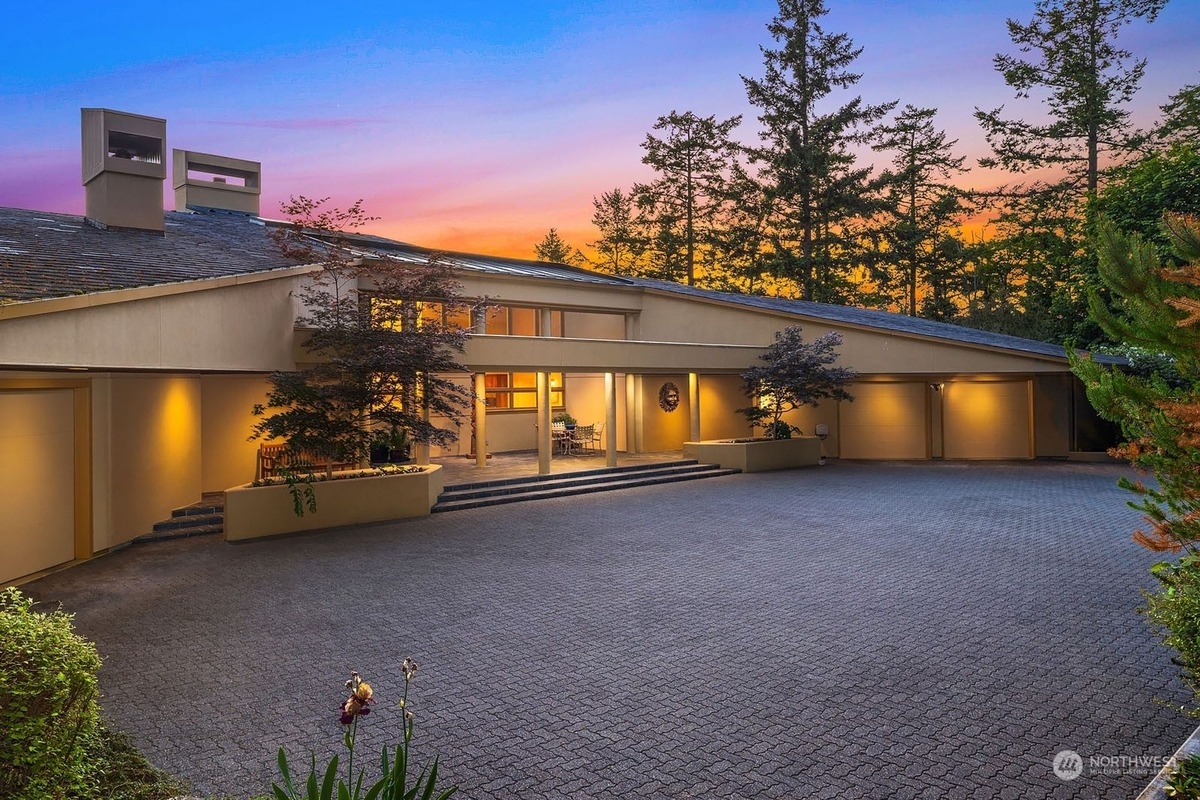 Large, contemporary home with a long driveway and expansive landscaping is illuminated at dusk.