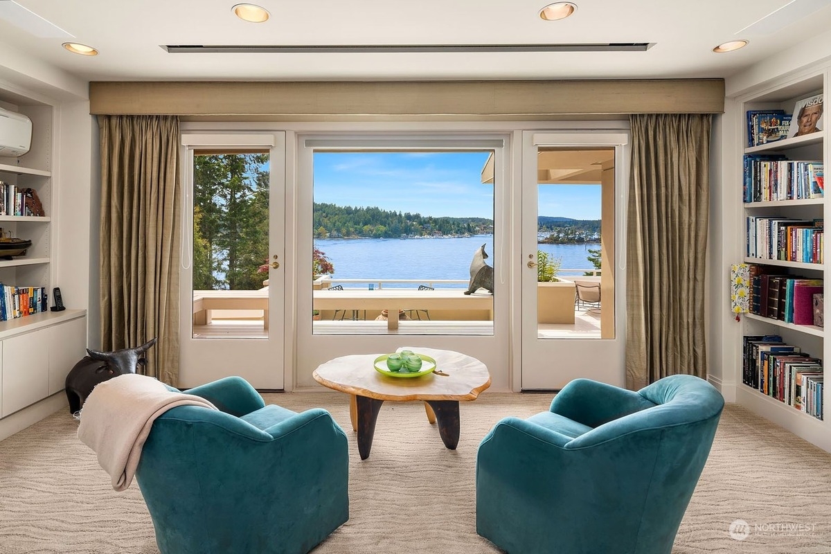 Room with teal armchairs and a wooden coffee table offers a stunning view of a lake and trees through large windows.