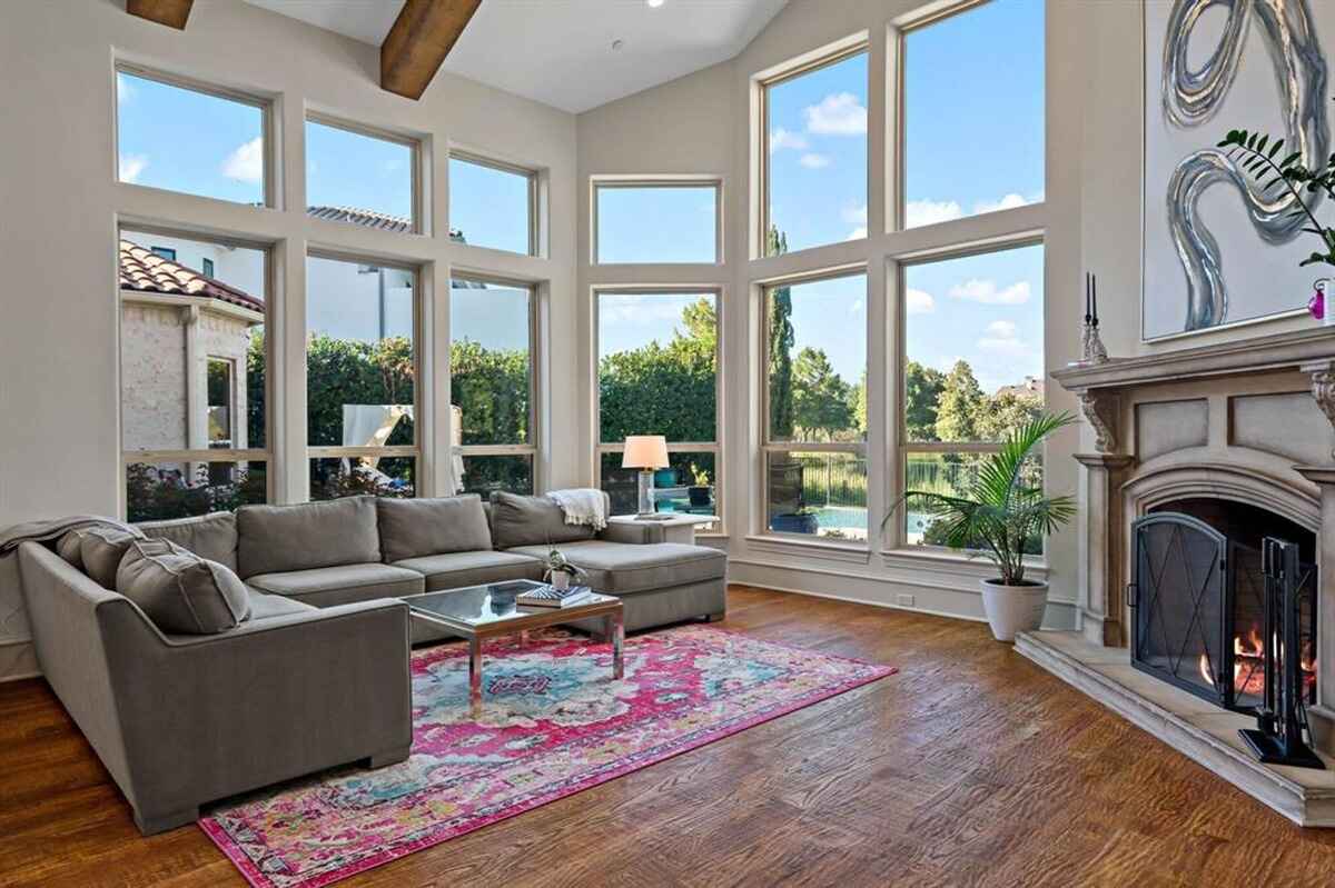 Living room includes large windows overlooking an outdoor view, a sectional sofa, a colorful area rug, a fireplace, and wooden floors with exposed ceiling beams.