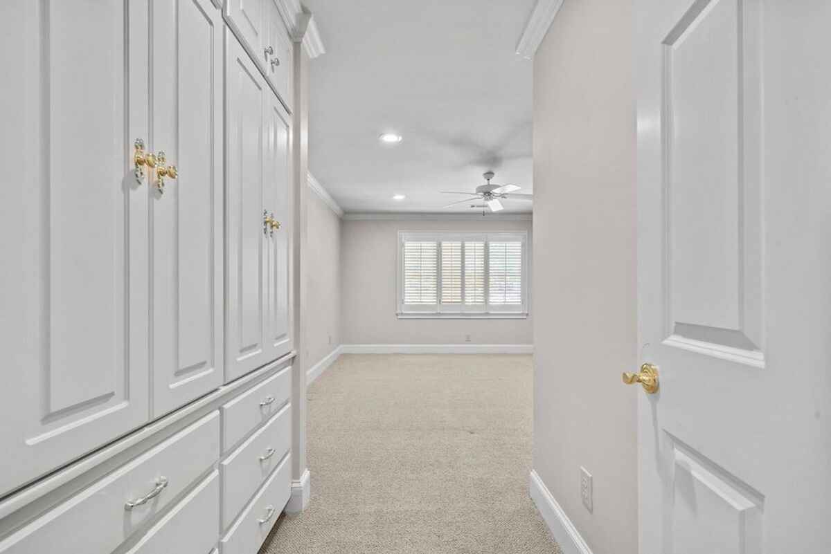 White built-in cabinets and drawers line one hallway wall; opposite, a carpeted bedroom with white walls and a ceiling fan is visible through an open doorway.