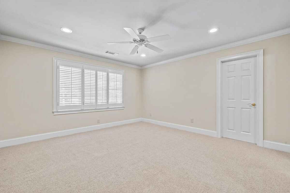 Carpeted bedroom with beige walls, white trim, window with shutters, and ceiling fan is shown.