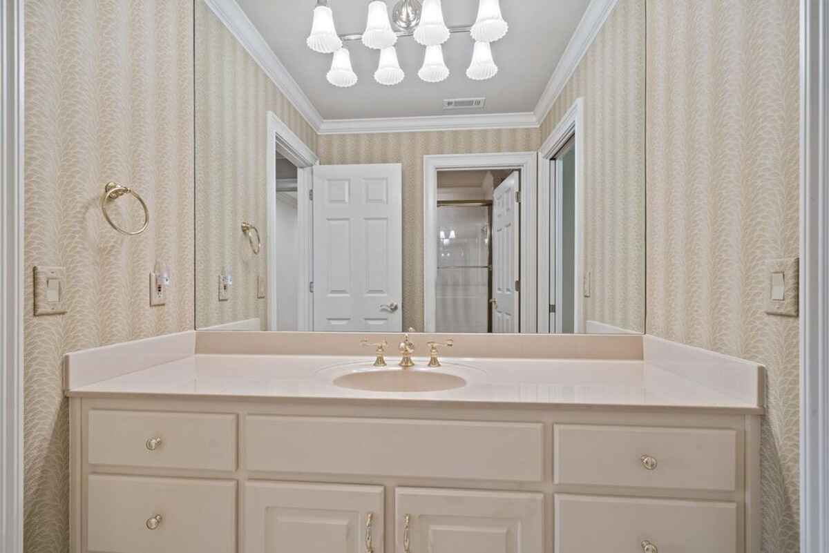 Off-white bathroom vanity with beige countertop, gold fixtures, and patterned wallpaper is shown.