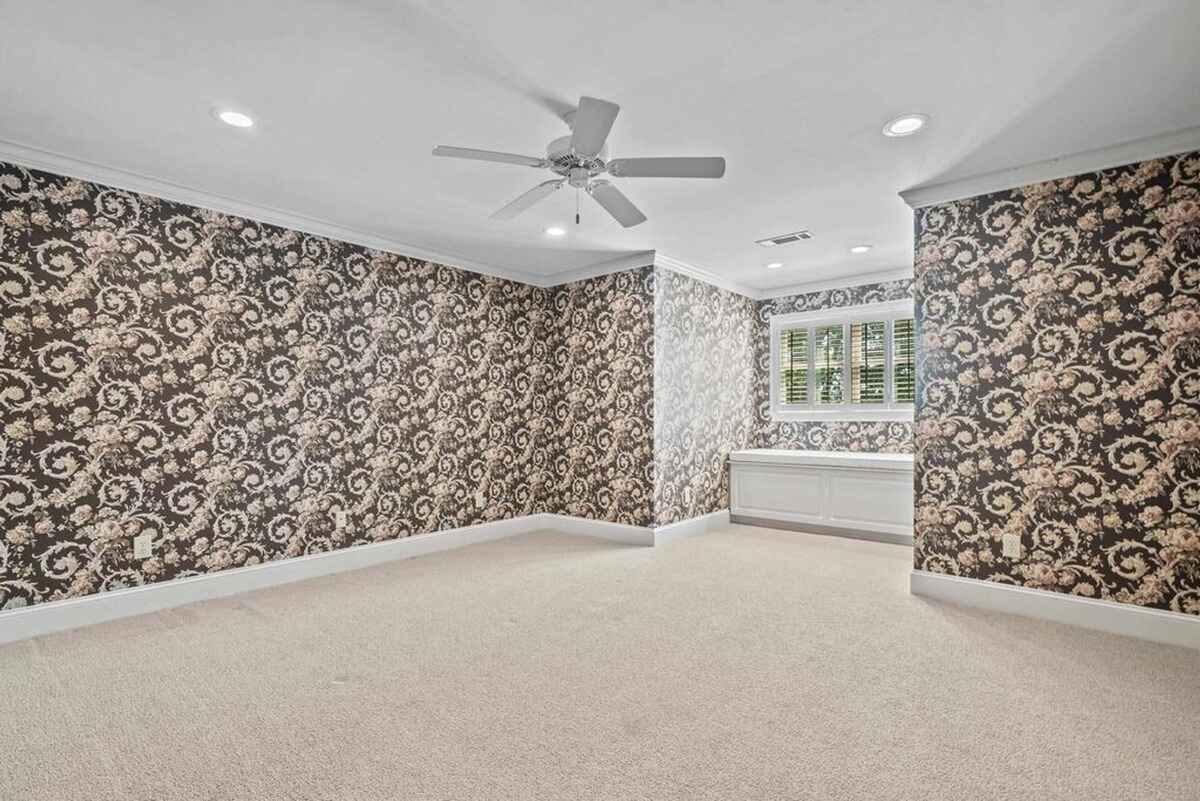 Carpeted room with patterned wallpaper, ceiling fan, and window with shutters.