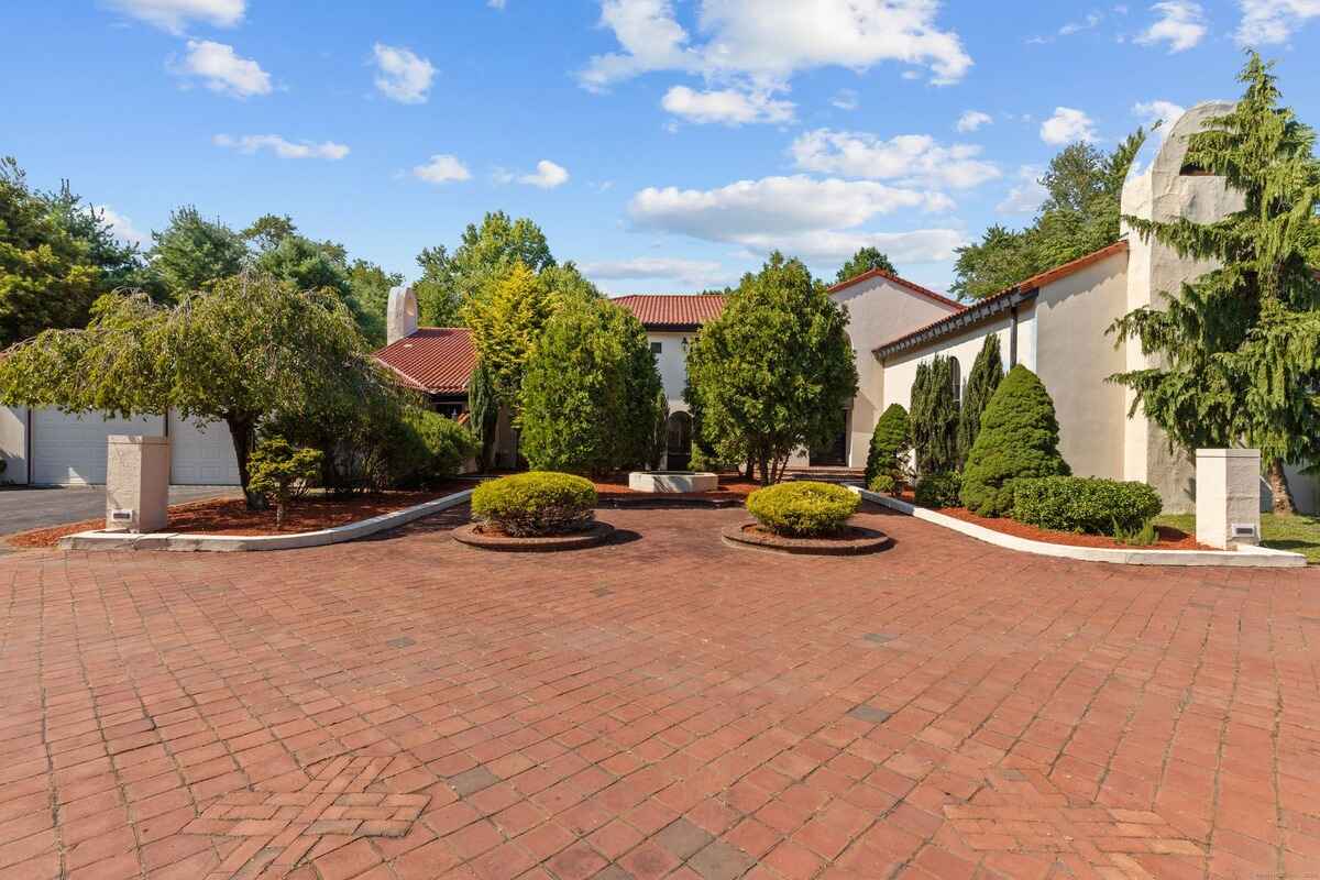 Brick driveway leading to a large, light-colored house with a red tile roof.