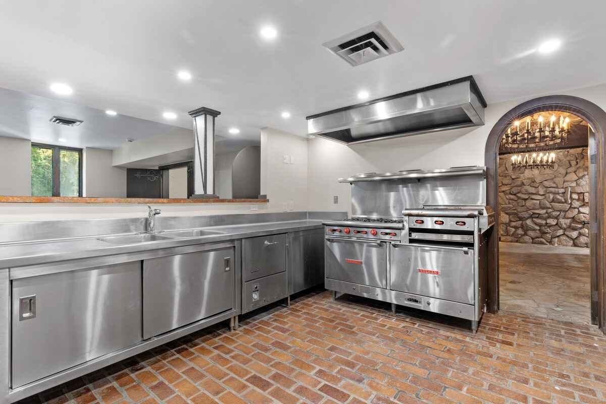 Commercial kitchen with stainless steel appliances and brick flooring, opening to a stone room.