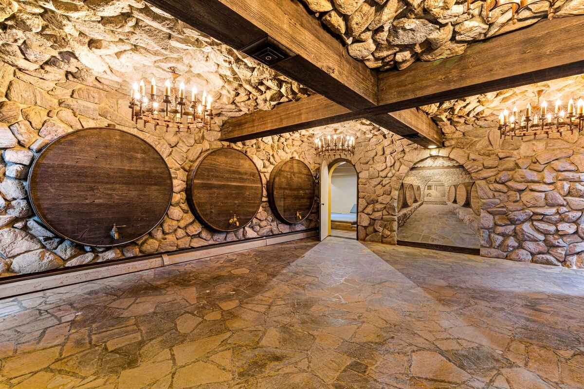 Wine cellar with stone walls, wood beams, large wooden barrels, and chandeliers.