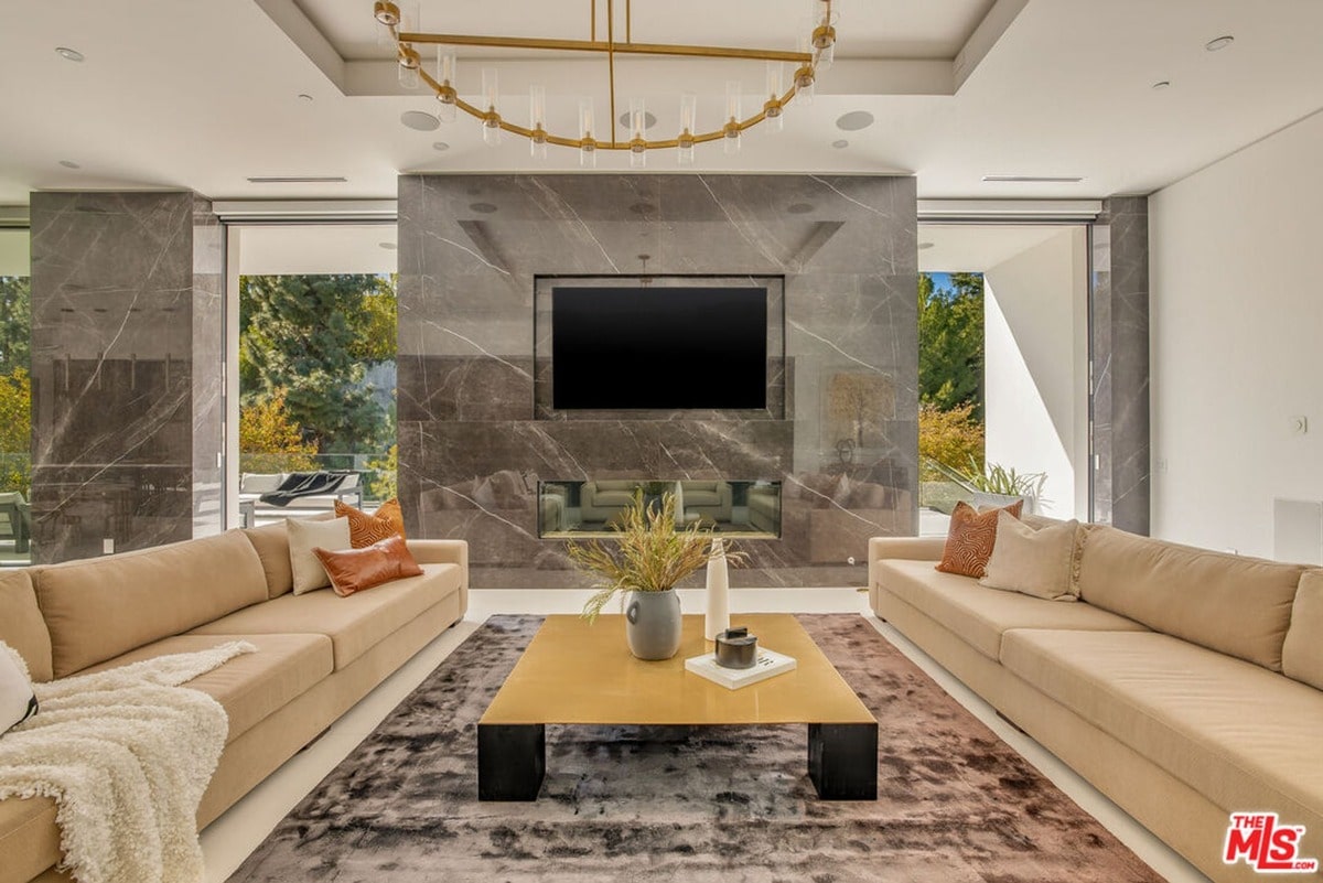 Elegance and understated luxury define this open-plan living area, where a large, modern fireplace commands attention. The room's neutral palette is warmed by natural light streaming through expansive windows, creating a relaxed and inviting atmosphere.