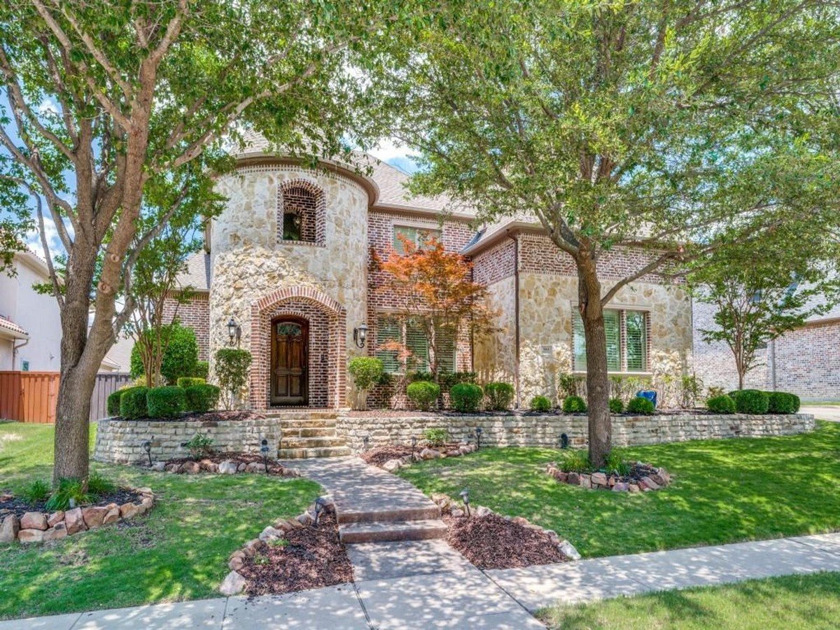 Two-story home with stone and brick exterior features a landscaped yard with mature trees.