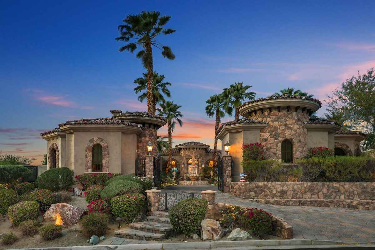 Front view of stone-accented house with circular towers, palm trees, and landscaped entrance.