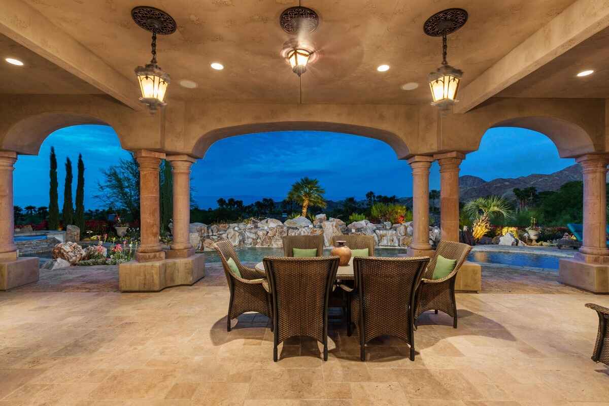 Covered patio with a dining table and lantern-style lights overlooking a landscaped pool area at dusk.