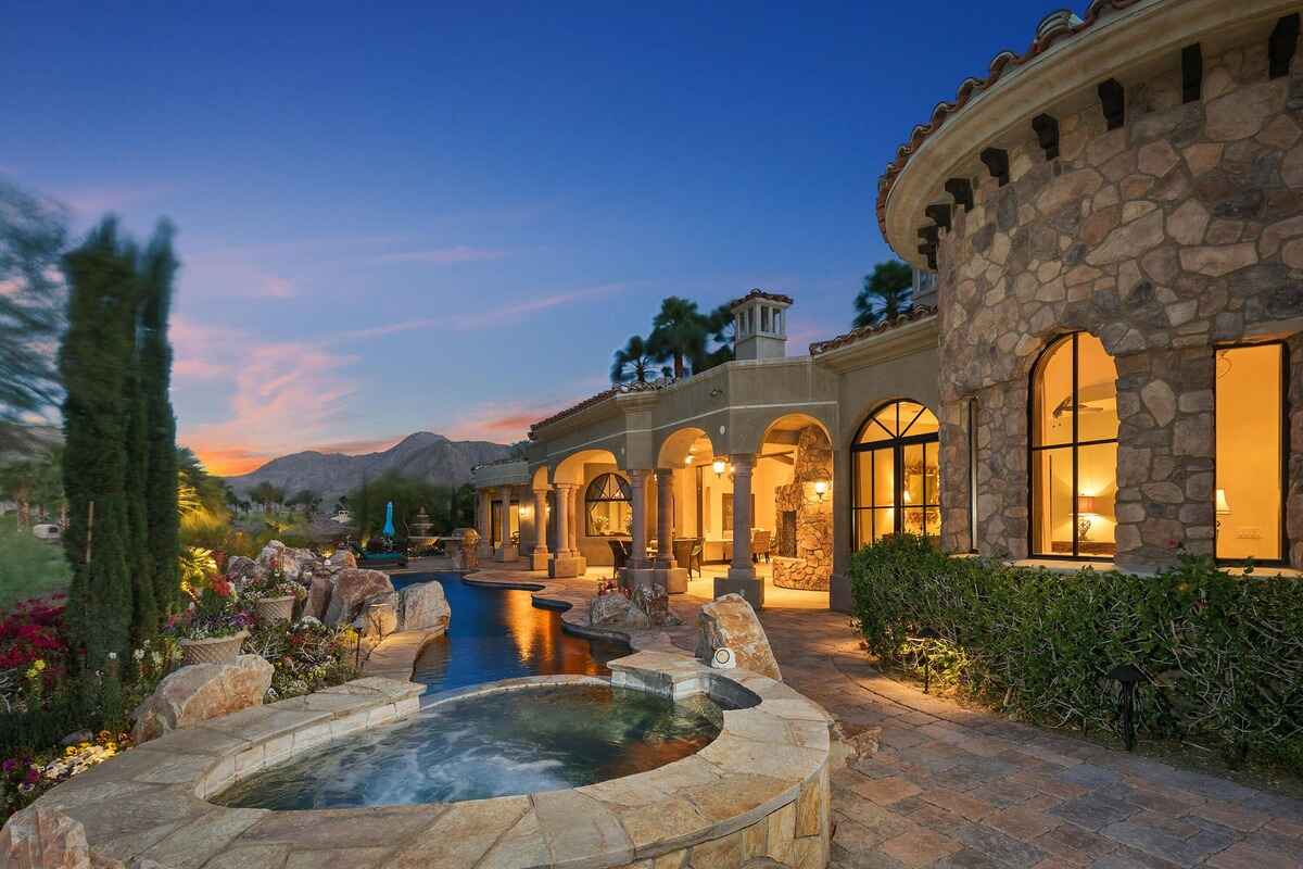 Exterior of a luxury home with a stone facade, outdoor spa, and mountain views under a twilight sky.