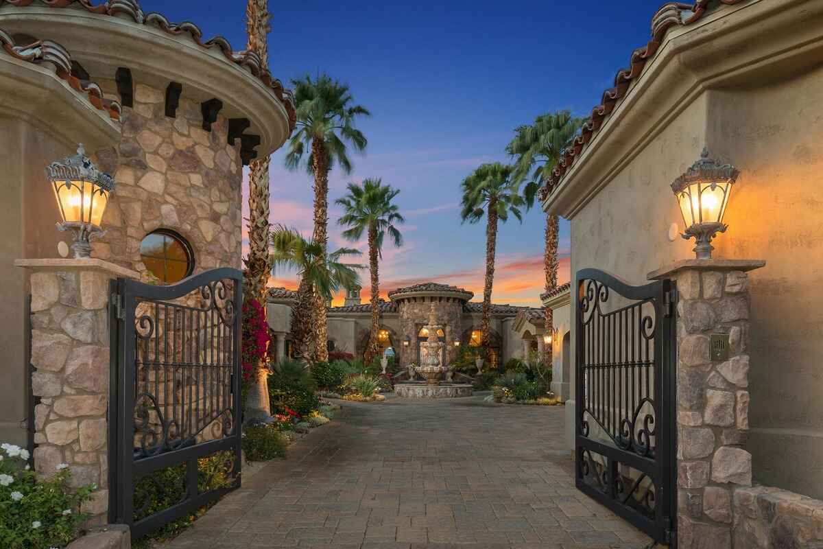 Elegant gated entry leading to a stone courtyard with lush landscaping and palm trees.