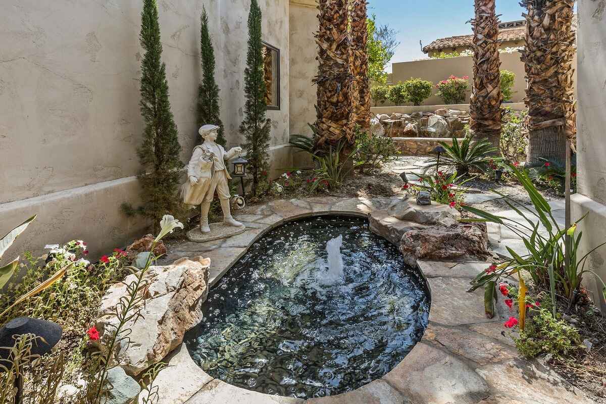 Garden walkway with small fountain, statues, and neatly arranged plants near palm trees.