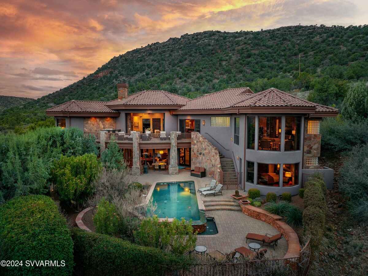 The image shows a Mediterranean-style home with a tiled roof, a pool, and surrounding greenery, set against a hillside backdrop at sunset.