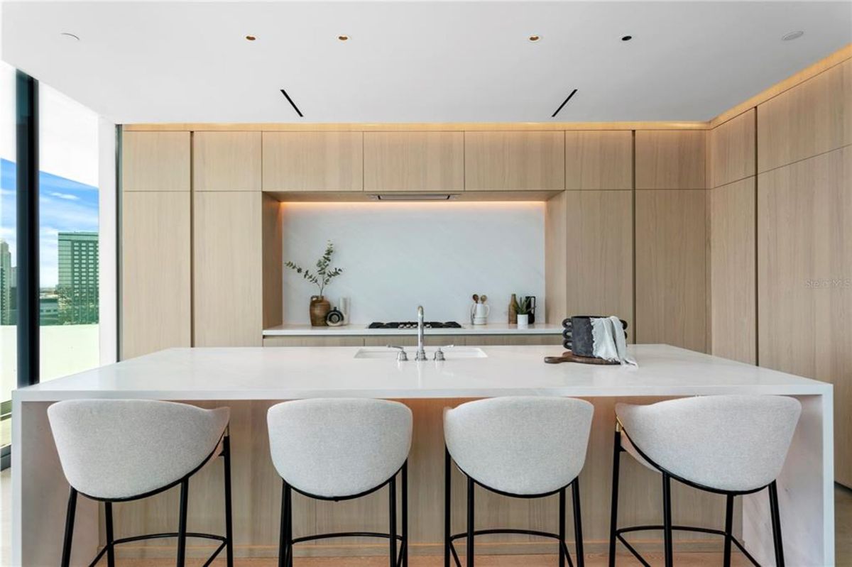 Kitchen with a large white island countertop that includes a built-in sink and four upholstered bar stools. Behind the island is a wall of light wood cabinetry with integrated appliances and a backsplash with soft lighting. Floor-to-ceiling windows provide natural light and offer views of nearby buildings and the cityscape.