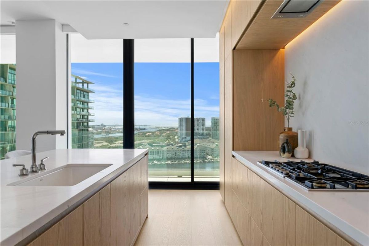 A modern kitchen with a large island featuring a white countertop and built-in sink. Adjacent to the island is a counter with a built-in gas cooktop, wooden cabinetry, and decorative items like vases with plants. Floor-to-ceiling windows provide views of nearby high-rise buildings and a distant waterfront under a clear sky.