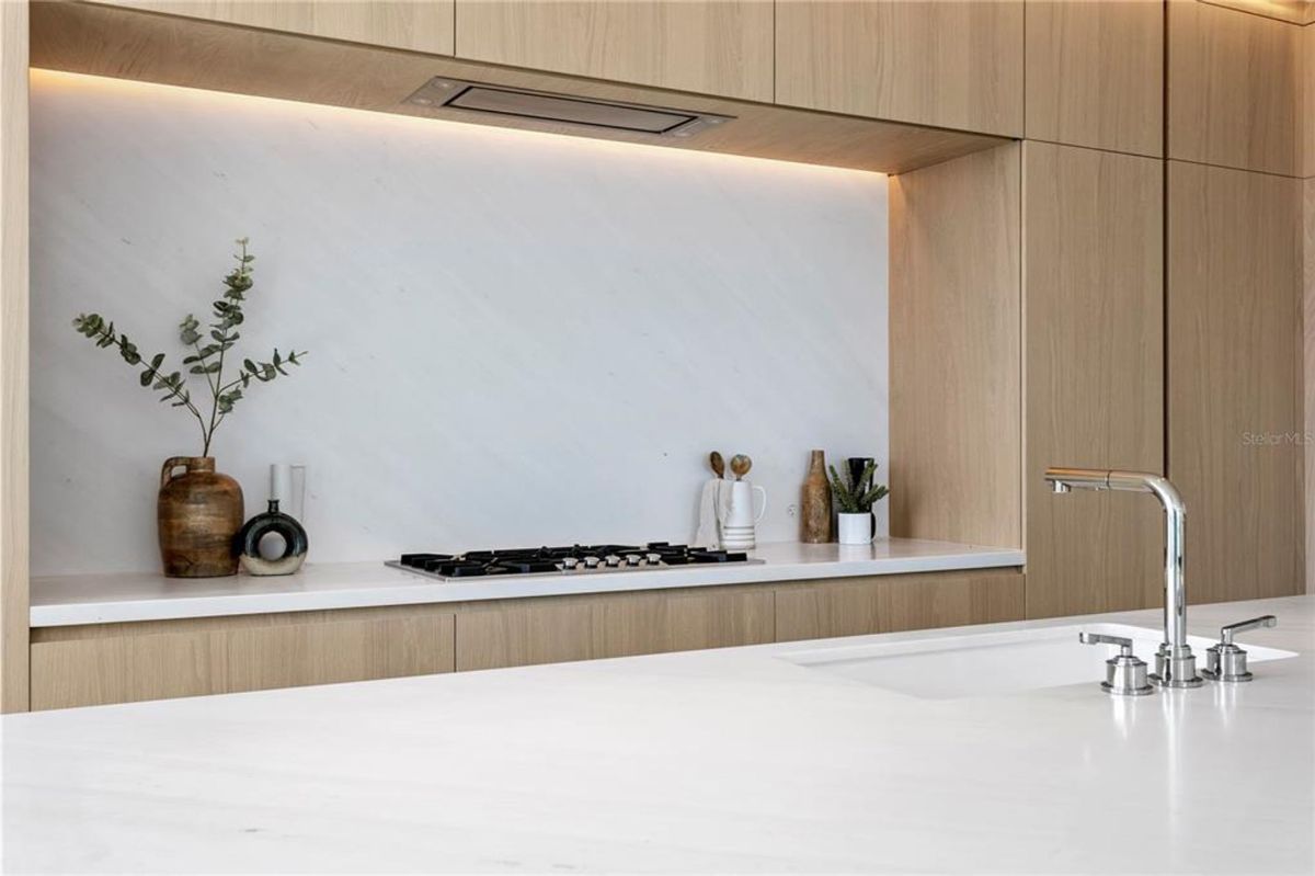 Kitchen with a built-in gas cooktop set into a white countertop, backed by a white marble backsplash. Above the cooktop, there is a wooden cabinet and an integrated vent hood. Decorative elements, such as potted plants and vases, are placed on the countertop, while a chrome faucet and sink are visible in the foreground.