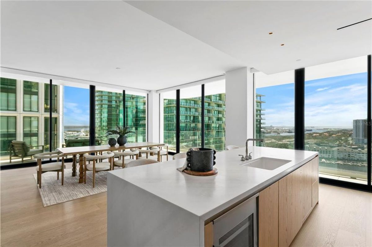 Kitchen island with a built-in sink, cabinetry, and a sleek countertop. A dining area with a wooden table and chairs is visible in the background, set near floor-to-ceiling windows. The windows provide views of surrounding high-rise buildings and a distant cityscape with clear skies.