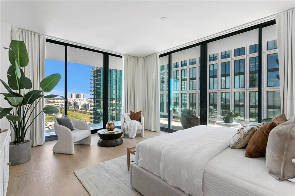 Modern bedroom with large floor-to-ceiling windows providing a view of nearby buildings and a blue sky. The room contains a bed with white linens and multiple pillows, two armchairs, a small side table, and a potted plant. A rug is placed under the bed, and long white curtains frame the windows.