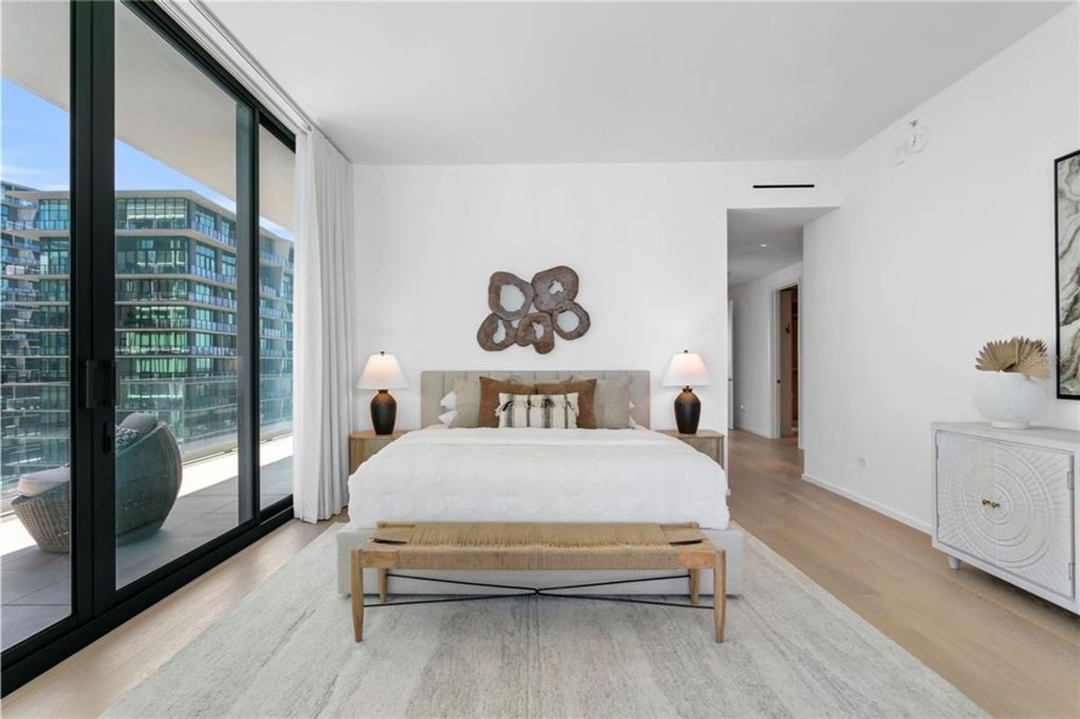 Bedroom with a large bed featuring white linens and multiple pillows. A decorative bench is positioned at the foot of the bed, with two bedside tables holding matching lamps on either side. Floor-to-ceiling windows reveal a balcony and adjacent buildings, while a white dresser and wall decor are visible in the corner of the room.