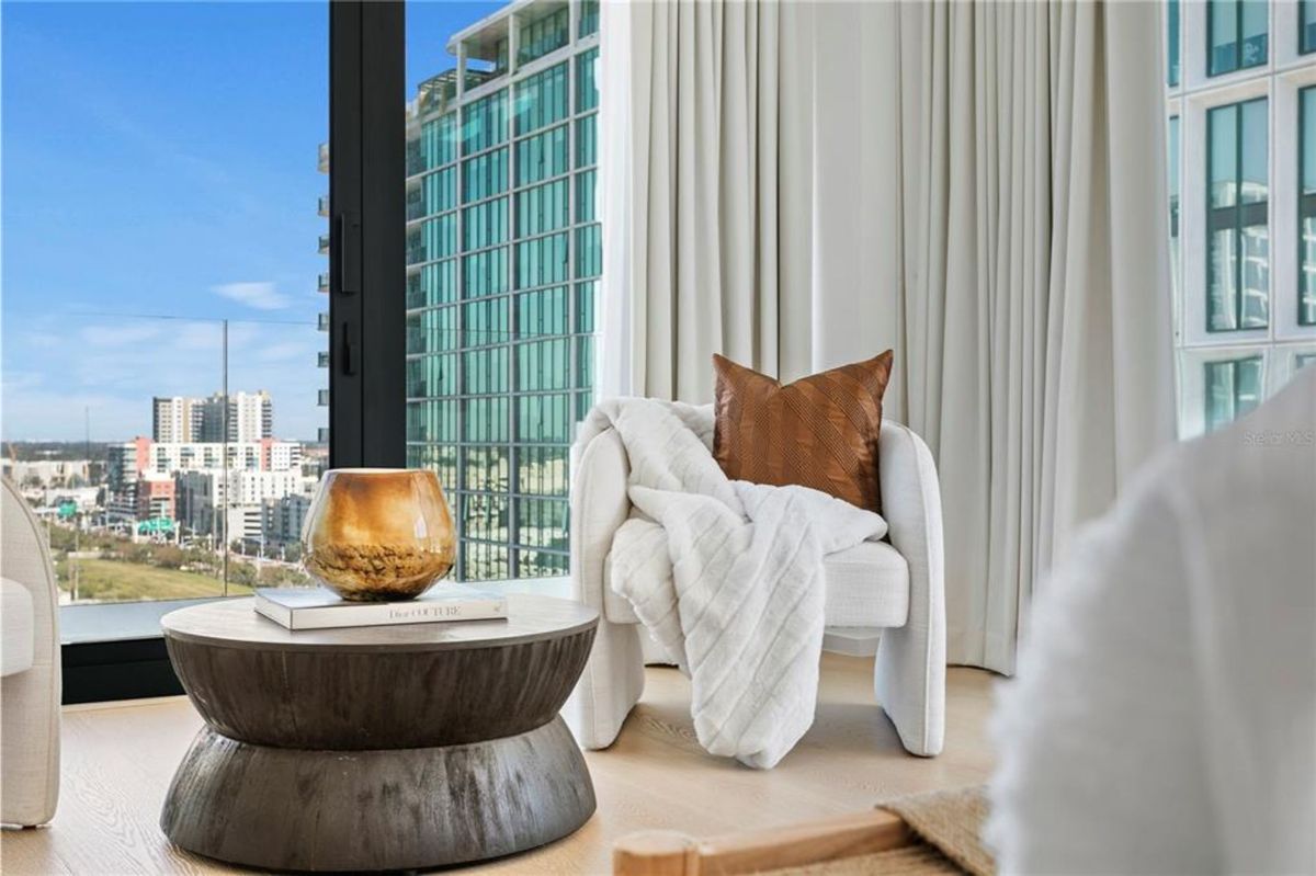 A cozy seating arrangement. The image shows a white upholstered armchair draped with a white blanket and accented with a brown throw pillow. A round dark wooden coffee table sits nearby with a decorative amber-toned glass object and books, while large floor-to-ceiling windows reveal an urban cityscape with high-rise buildings.